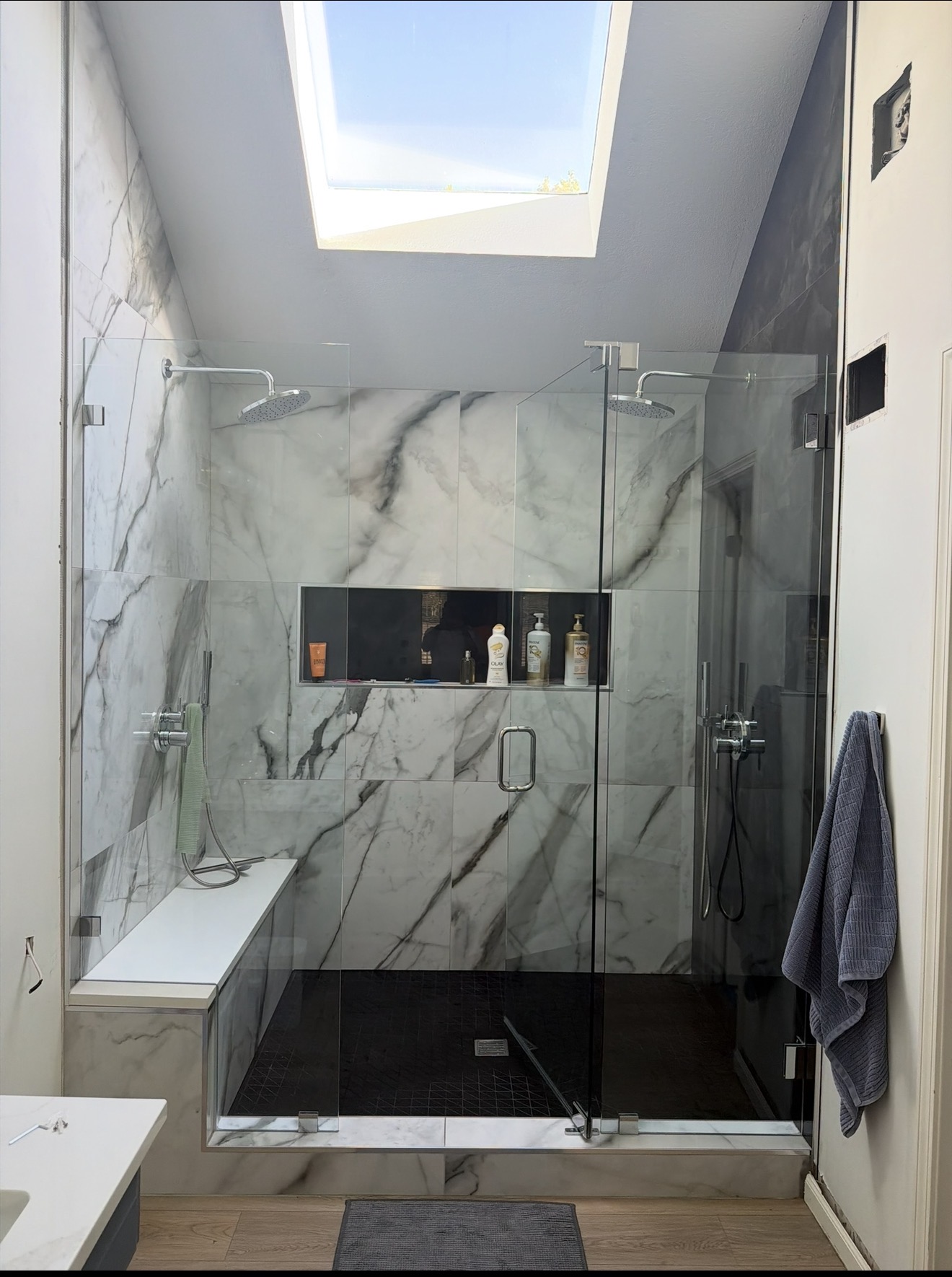 Completed large glass-enclosed shower in marble-look tile with two rain heads, a full-width niche and a built-in bench, beneath the existing skylight