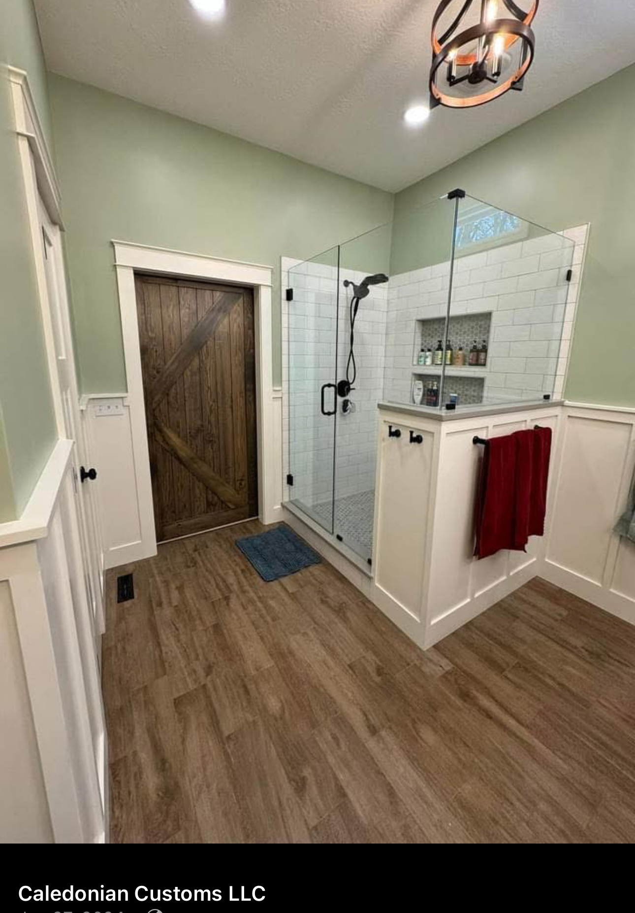 Completed primary bathroom with a glass shower, rustic wood door, sage-green walls, white wainscot and red towel