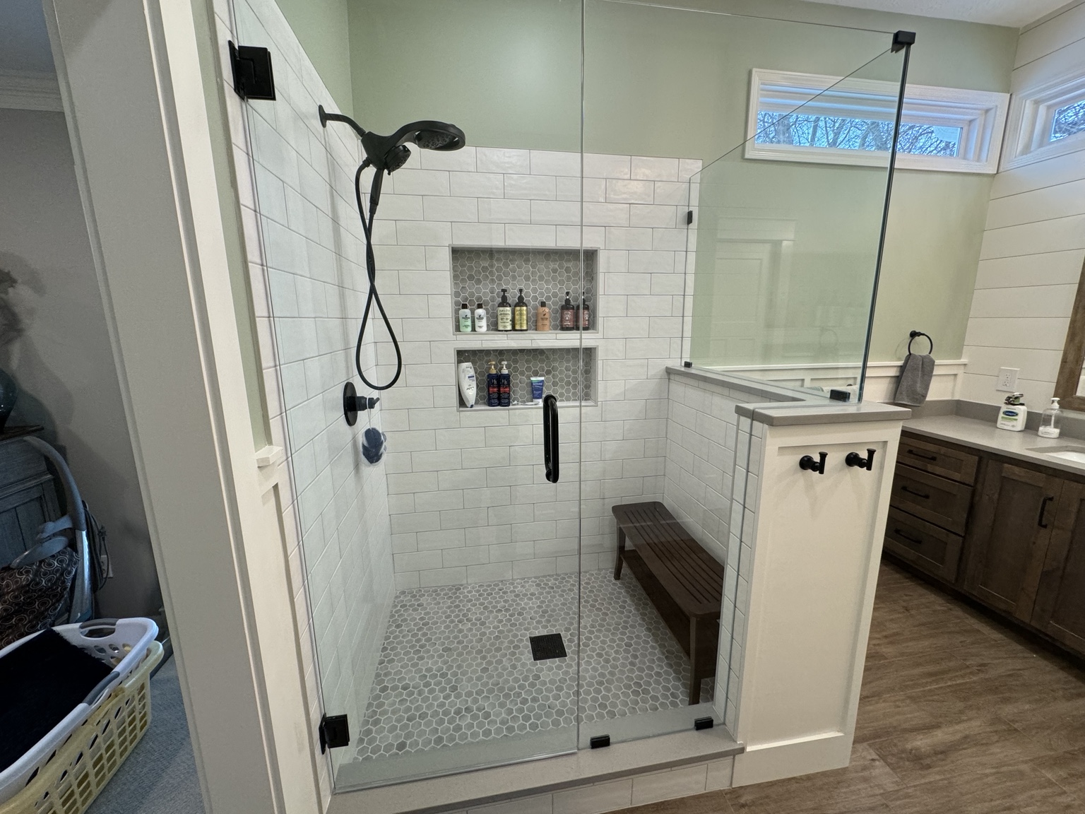 Completed custom glass shower with subway tile, hexagon mosaic floor, two niches and a teak bench, with the wood vanity beyond