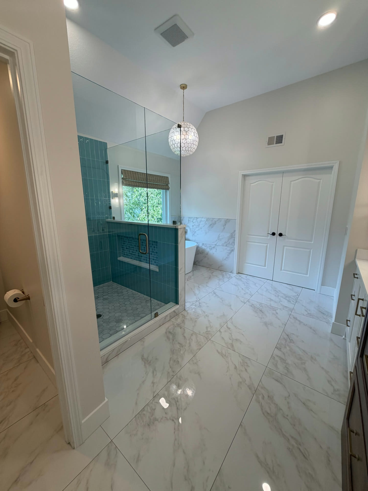 Completed primary bathroom looking across the marble-look tile floor toward the glass shower and double closet doors