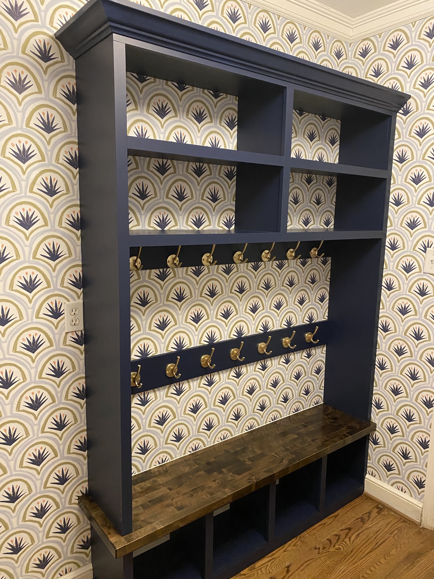 Completed navy mudroom built-in with upper cubbies, two rows of brass hooks and a stained wood bench