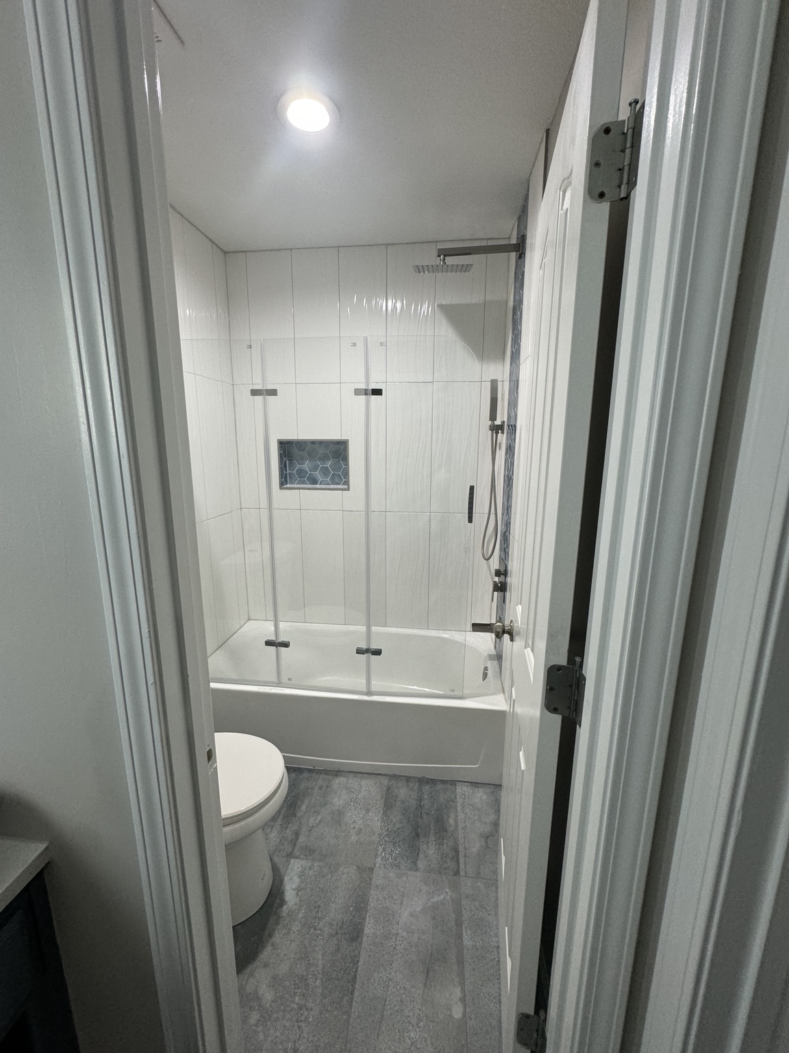 Completed tub and shower seen through the doorway, with white textured wall tile, a glass enclosure and grey wood-look floor tile
