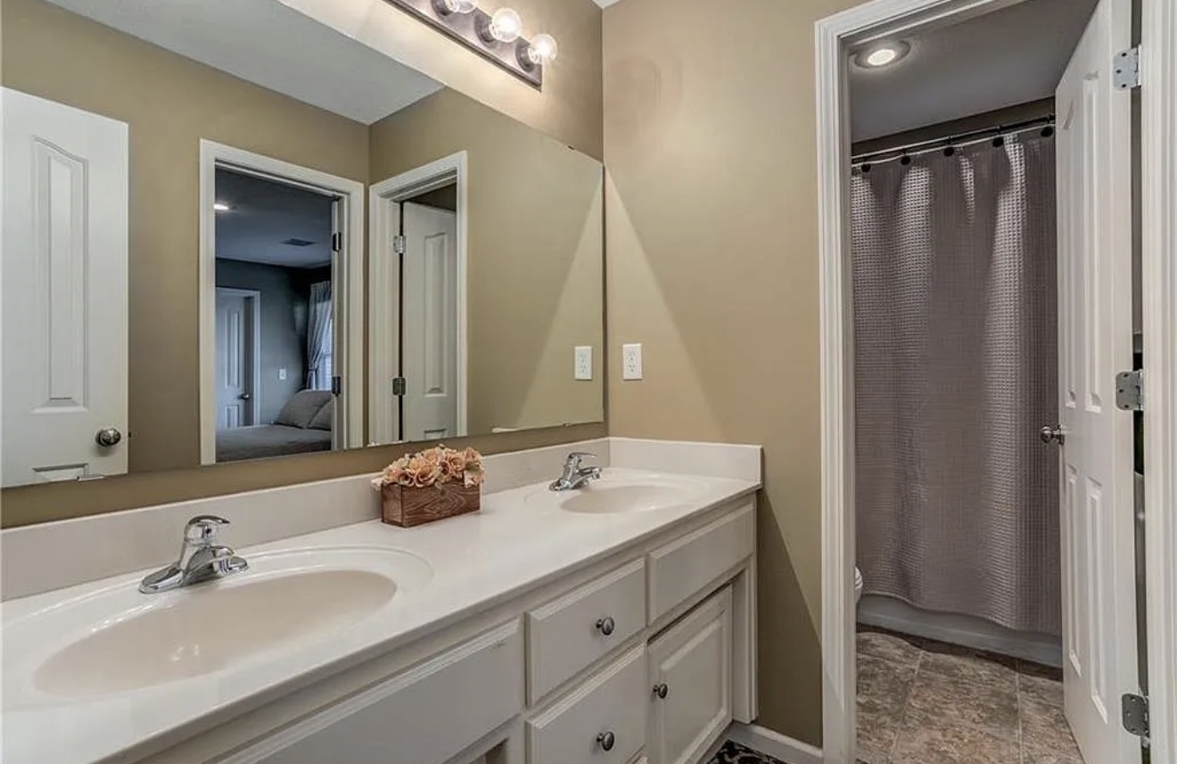 Original bathroom with tan walls, a long white vanity with two oval sinks, a frameless mirror and a curtained tub area beyond the doorway