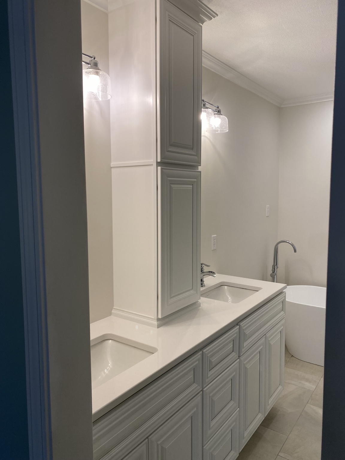 Finished double vanity with two undermount sinks, a white quartz-look countertop, grey raised-panel cabinetry, a tall centre tower cabinet, mirrors and seeded-glass sconces, with the freestanding tub beyond