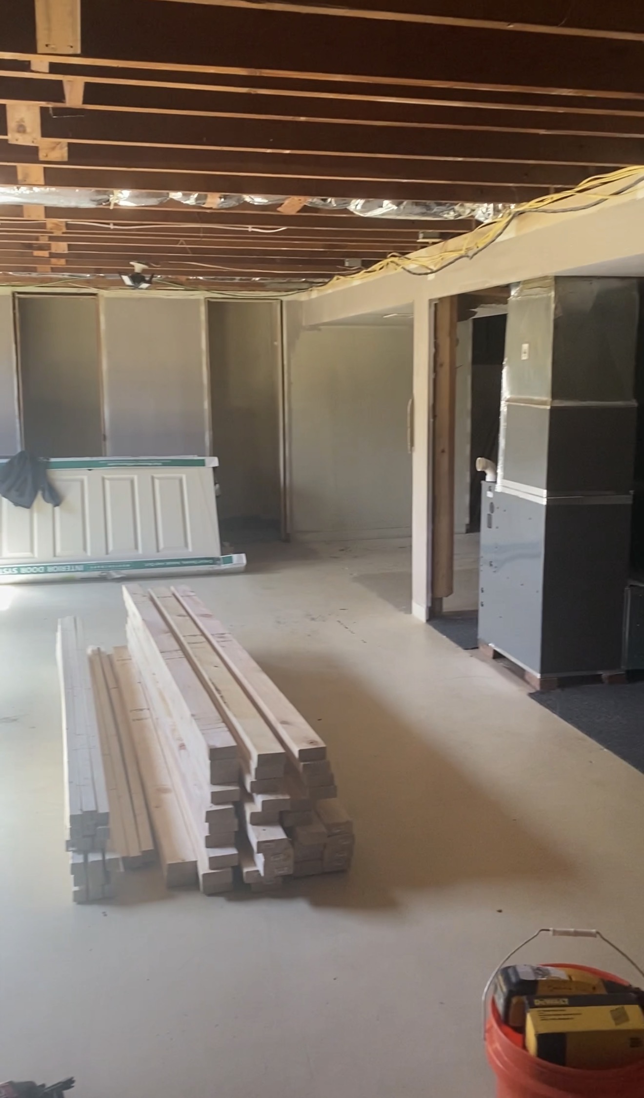 The same basement mid-construction, with exposed ceiling joists, wiring and ductwork above, a stack of framing lumber on the floor and a new door still in its packaging