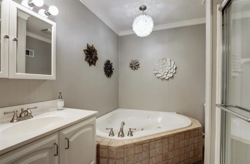 The original bathroom with a corner jetted tub set in tan tile, grey walls, a white vanity with a framed mirror and an enclosed shower to the right