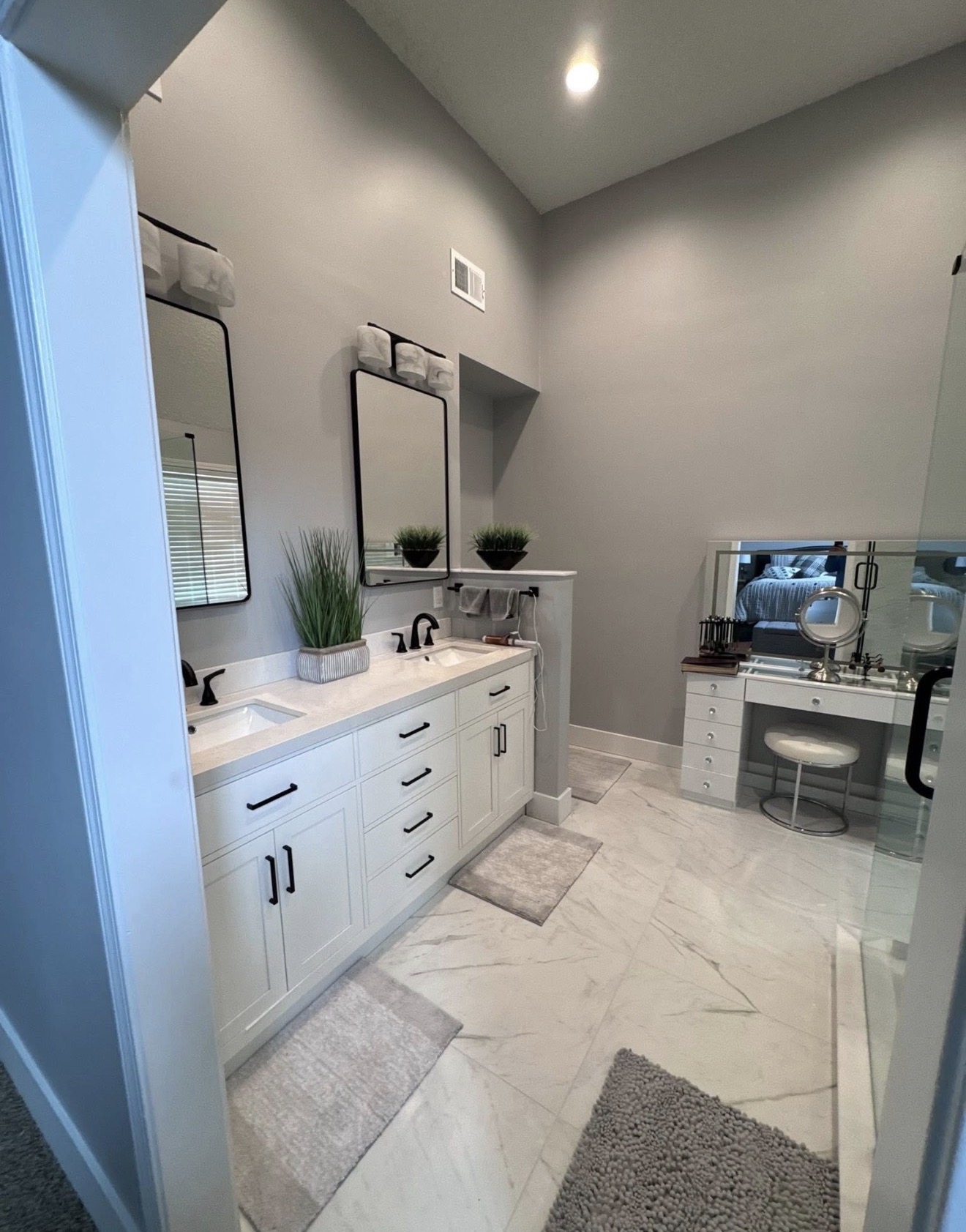 Completed double vanity with white cabinetry, black hardware, a makeup station and the bedroom visible in the background