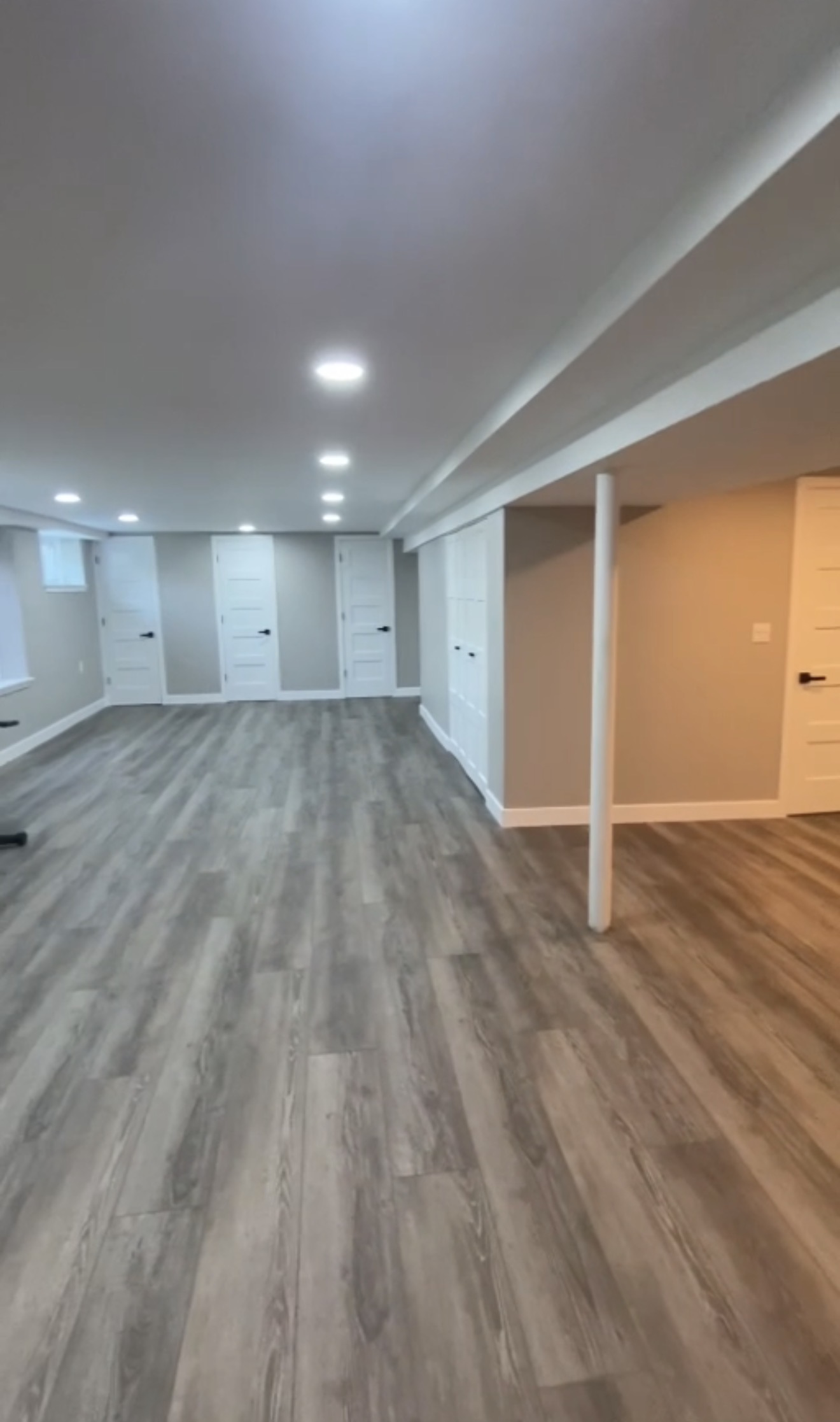Completed open basement living space with grey painted walls, a tan accent wall, wood-look plank flooring, white doors and trim, recessed ceiling lighting and a white-wrapped support post