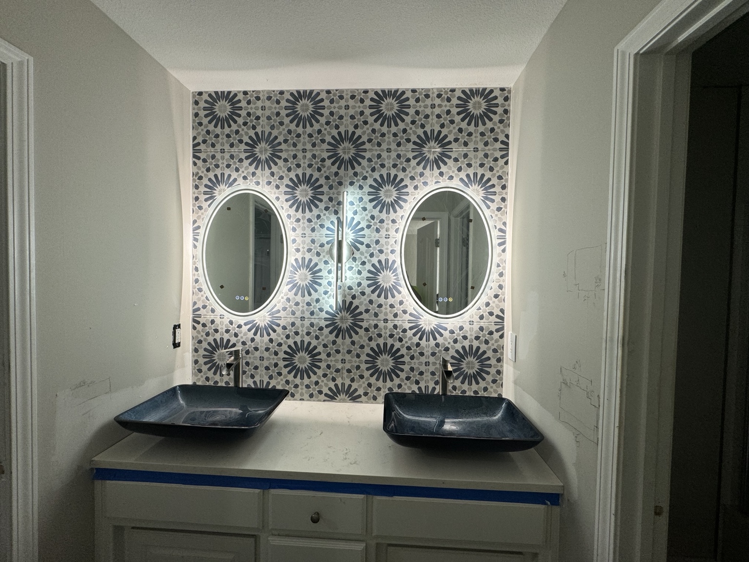 Completed vanity wall tiled floor to ceiling in a blue and white patterned tile behind two oval lighted mirrors and two blue vessel sinks