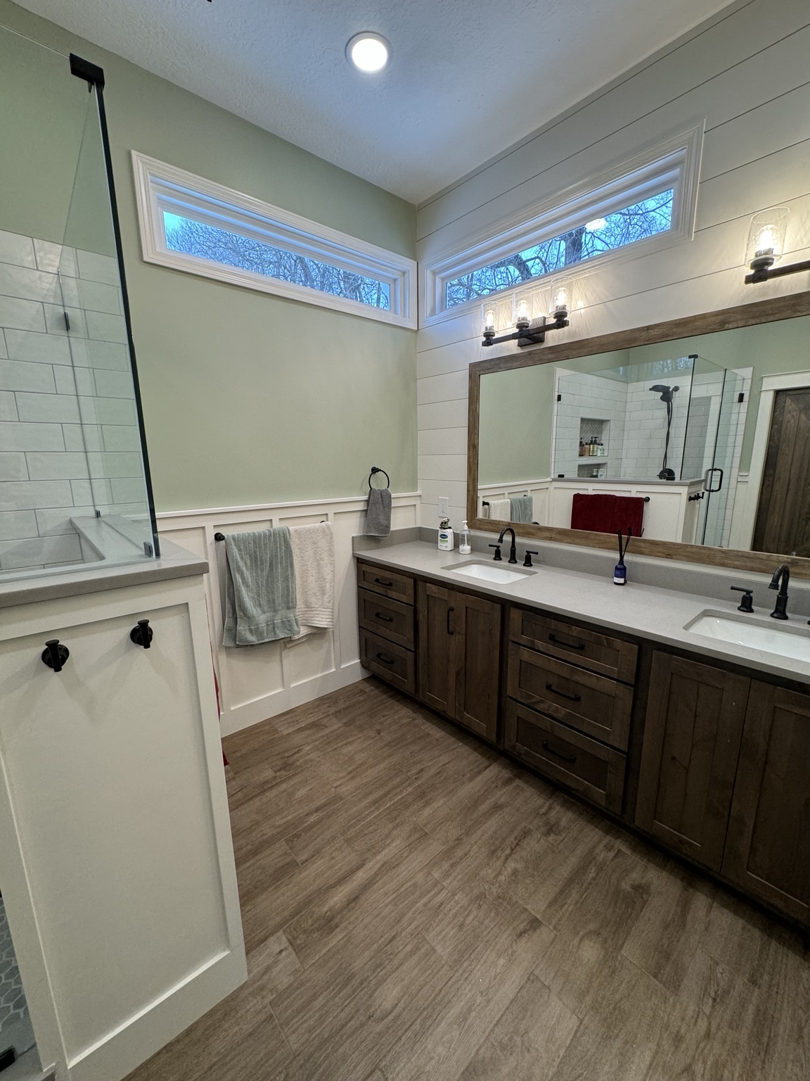 Completed double vanity with wood cabinetry, framed mirror, sage-green walls, white wainscot and transom windows