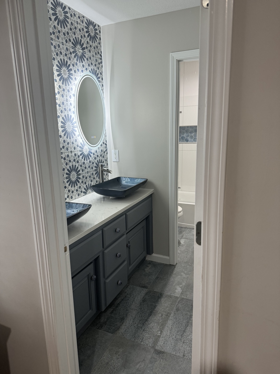 Completed bathroom from the hallway, showing the grey vanity with blue vessel sinks, patterned tile wall and the tub beyond