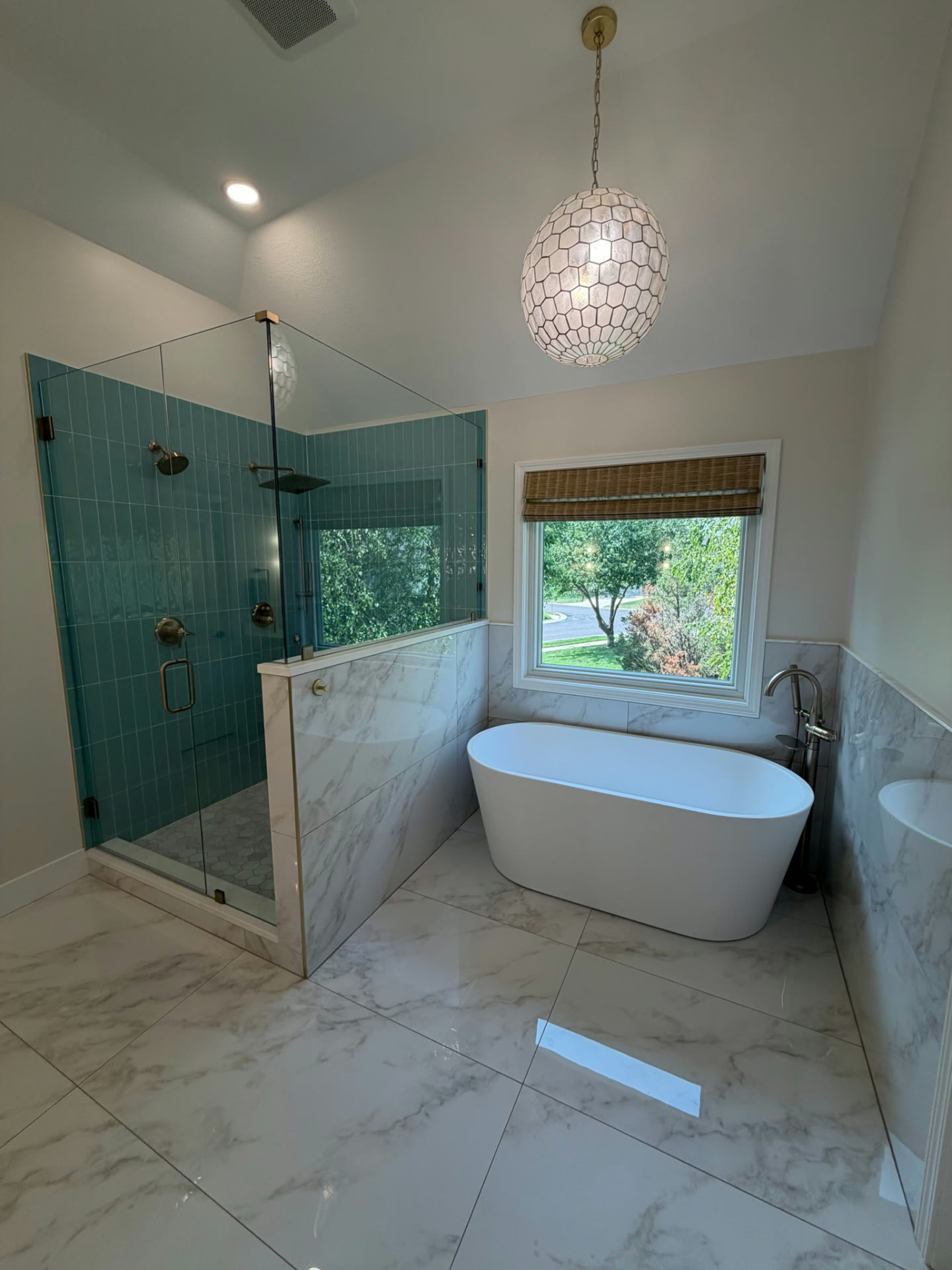 Completed primary bathroom with teal vertical-tile glass shower, freestanding tub beneath the window and a globe pendant light