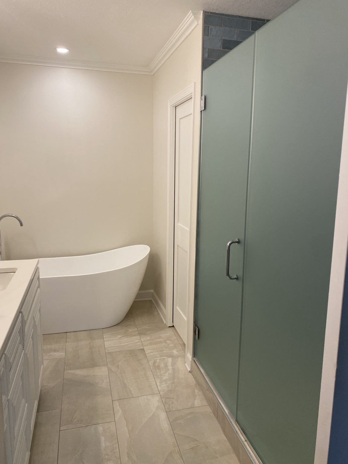 Completed primary bathroom with a white freestanding tub against a cream wall, a glass-doored custom shower to the right, large marble-look floor tile, white crown moulding and a recessed ceiling light