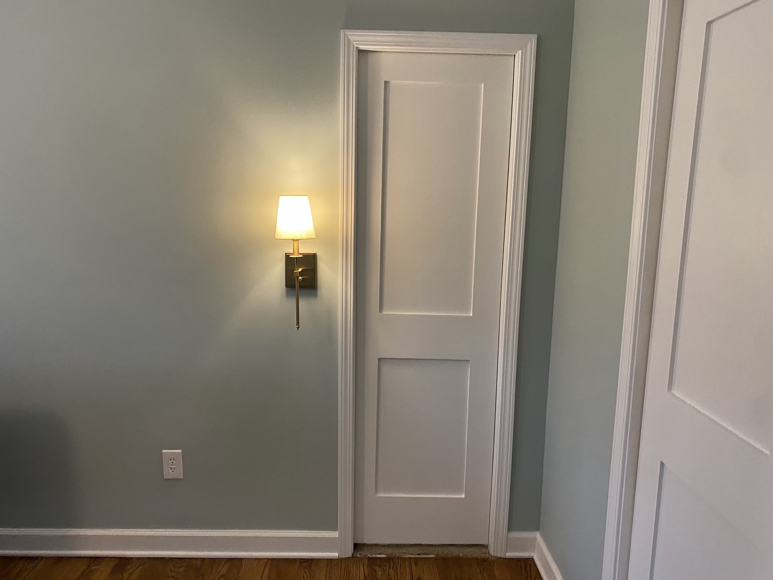 Close view of the new white pocket door in the bedroom wall, with fresh casing, a brass wall sconce alongside and hardwood flooring below